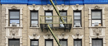 The facade of a residential prewar brick building with a green fire escape and a bright blue cornice in West Harlem, Manhattan, New York City